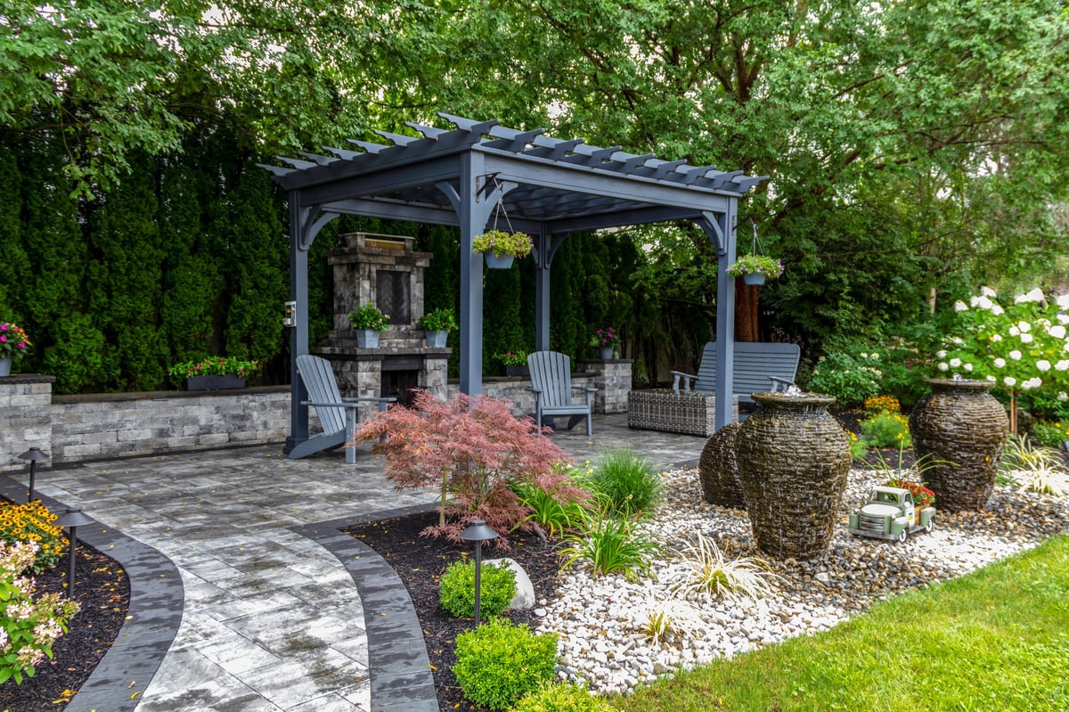 Large gray pergola on pavers with stone fireplace and waterfall urns for backyard oasis landscape architecture