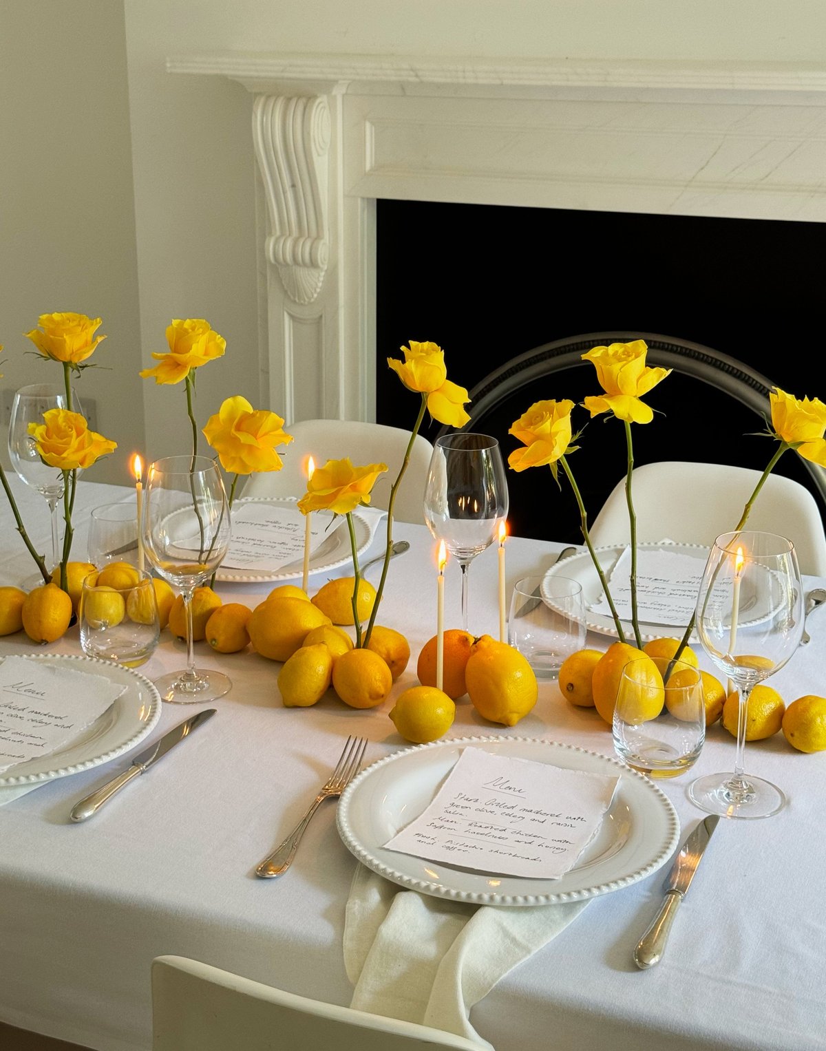 A beautifully arranged summer tablescape featuring bright yellow roses and fresh lemons scattered across a crisp white tablecloth