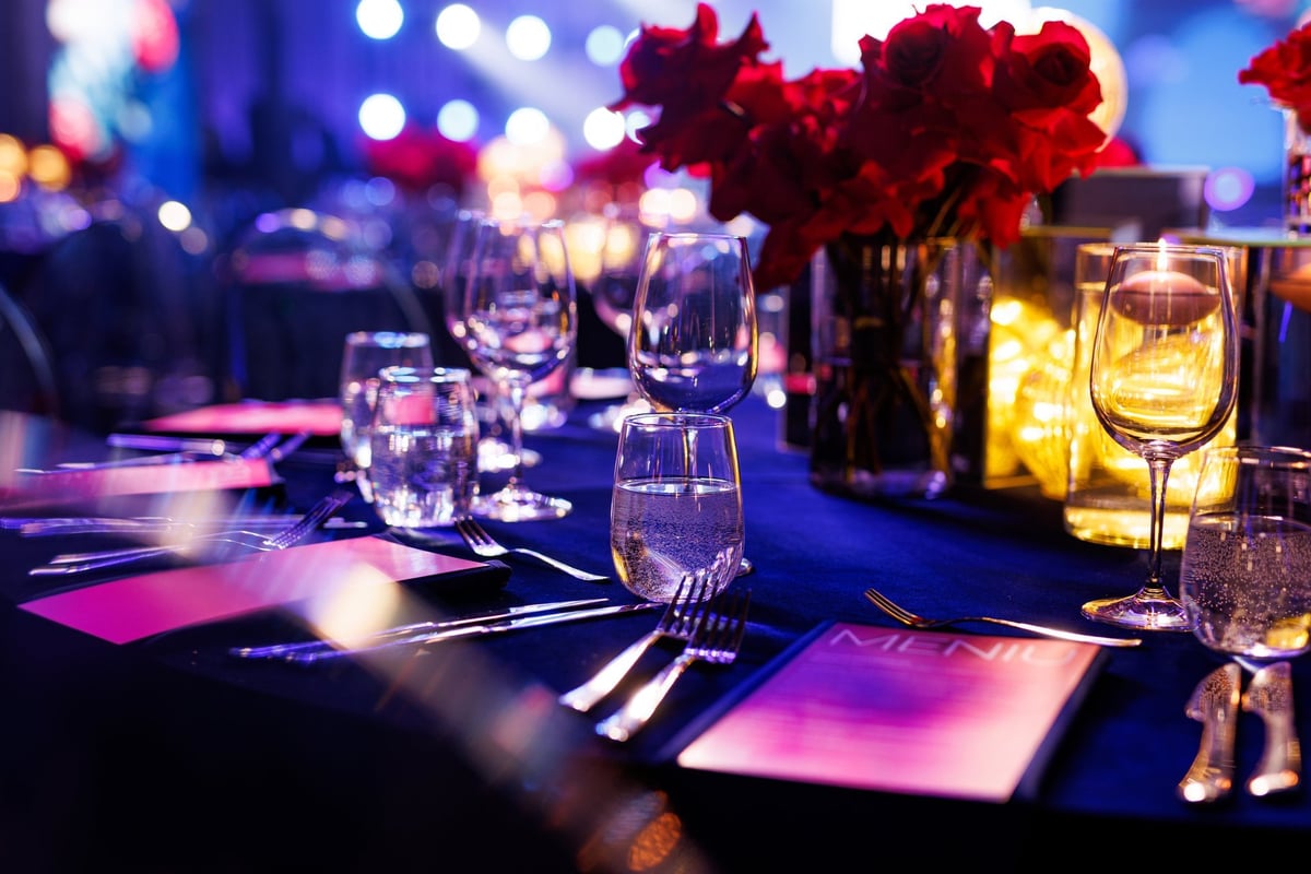 A formally set banquet table with glassware, menus, and floral centerpieces under blue atmospheric lighting